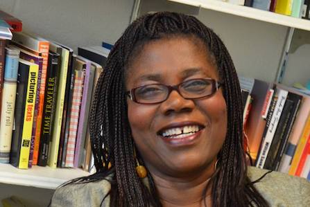 A black woman with braided hair and glasses smiling in front of a bookshelf.